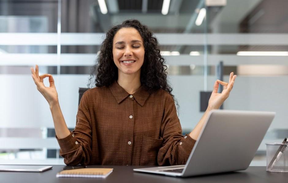 Mulher meditando com olhos fechados e sorrindo à mesa do escritório, transmitindo tranquilidade e equilíbrio no trabalho.