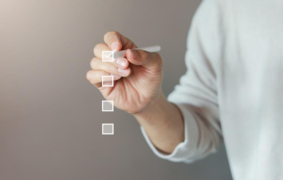 Mão marcando o primeiro item de uma checklist virtual, simbolizando tarefas ou avaliação.