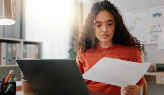 Jovem profissional analisa um documento com atenção em seu home office, com laptop aberto à frente e um mural de referências ao fundo.