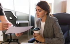 Mulher em ambiente corporativo avaliando um documento enquanto segura um café, com postura focada e analítica.