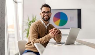 Homem sorridente de negócios em ambiente de escritório, com laptop e gráficos de análise de dados ao fundo, enfatizando sucesso e produtividade.