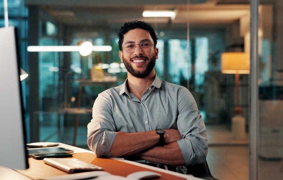 Homem jovem com óculos e barba, sorrindo com os braços cruzados, sentado em uma mesa de escritório moderno com iluminação noturna ao fundo.