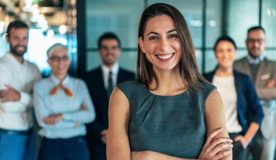  Mulher sorridente em traje formal cinza com braços cruzados, em destaque à frente de sua equipe.