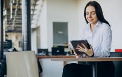 Uma mulher sorridente em trajes executivos sentada à mesa de um escritório moderno, focada enquanto utiliza um tablet.