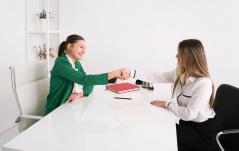 Duas mulheres se cumprimentam com aperto de mão através de uma mesa branca, sugerindo uma entrevista de emprego ou reunião profissional.