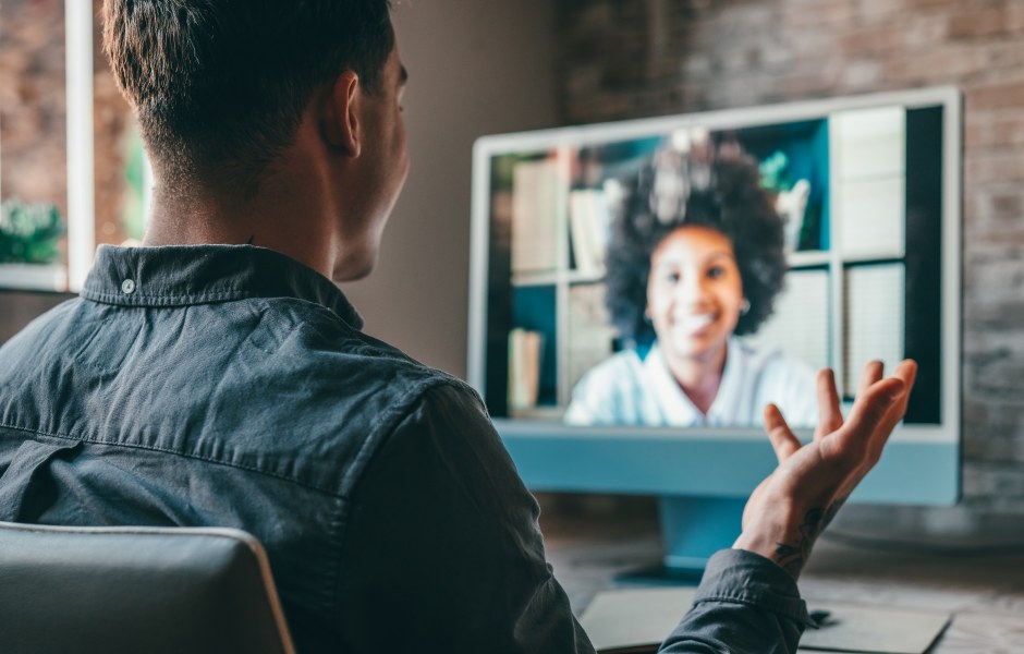 Homem em videochamada gesticulando para colega que sorri na tela do computador, representando uma reunião ou entrevista remota.
