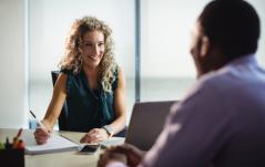 Mulher sorridente anota em caderno enquanto conversa com um colega, sugerindo uma reunião one-on-one ou entrevista profissional.