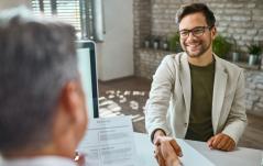 Homem de óculos sorri e aperta a mão do entrevistador, com currículo sobre a mesa, em uma entrevista de emprego.