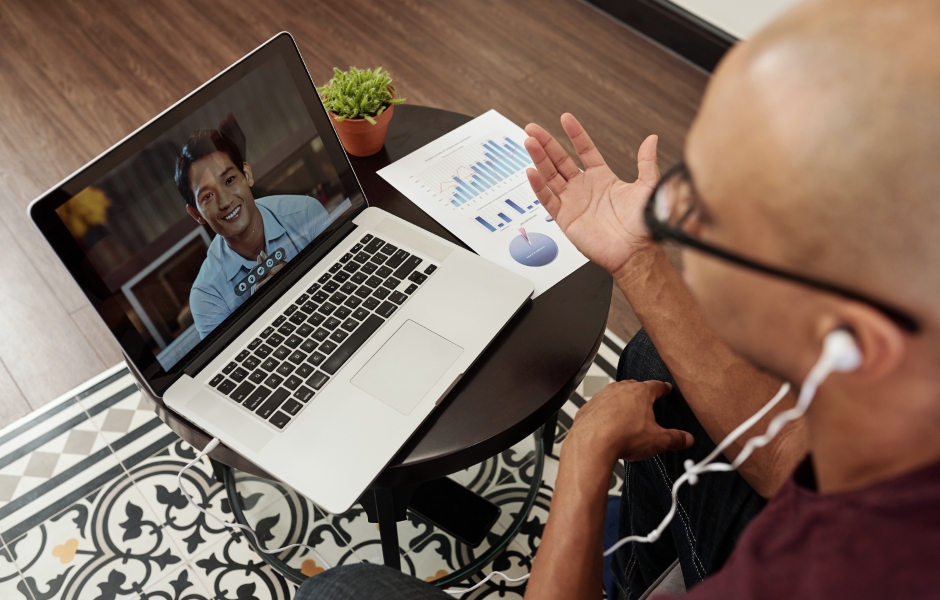 Homem fazendo videoconferência em um laptop, interagindo com outra pessoa na tela, com gráficos ao lado.