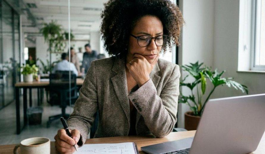 Mulher de óculos observa a tela do notebook com a mão no queixo em tom concentrado. Ela toma notas em um caderno ao lado de uma caneca sobre a mesa de madeira. Ao fundo, vê-se um escritório moderno, bem iluminado e decorado com plantas.