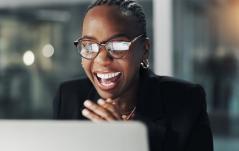 Mulher corporativa celebrando e sorrindo enquanto trabalha no computador, usando óculos e roupa elegante, representando sucesso, inovação e profissionalismo.