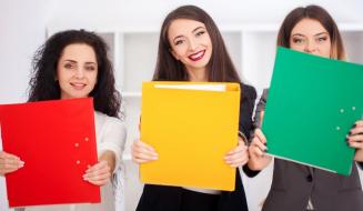 Três mulheres sorridentes seguram pastas coloridas (vermelha, amarela e verde) à frente do corpo em um ambiente de escritório.