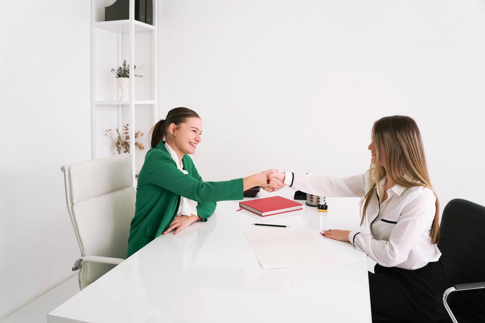 Duas mulheres se cumprimentam com aperto de mão através de uma mesa branca, sugerindo uma entrevista de emprego ou reunião profissional.