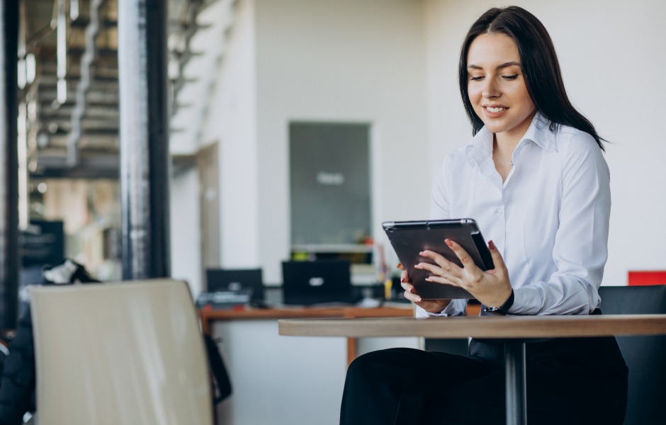 Uma mulher sorridente em trajes executivos sentada à mesa de um escritório moderno, focada enquanto utiliza um tablet.
