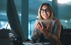 Executiva de óculos sorri segurando um tablet em escritório iluminado com luz azulada noturna.