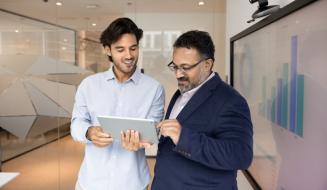 Dois homens de traje social discutem dados exibidos em um tablet em ambiente de escritório moderno, promovendo comunicação eficiente e análise de negócios.