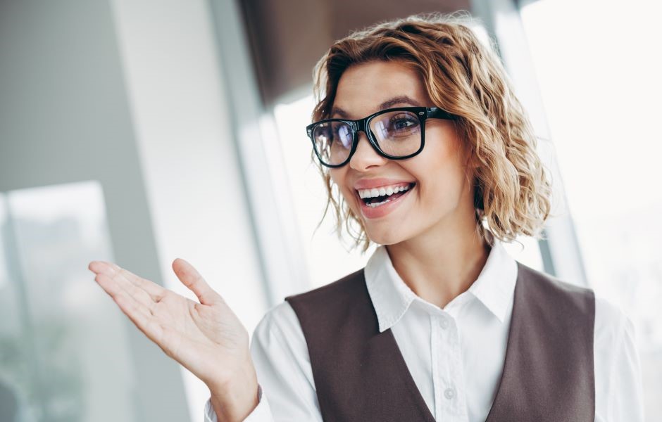 Mulher empresária sorridente usando óculos de grau, vestindo camisa branca e colete, em ambiente de escritório, demonstrando entusiasmo e confiança.