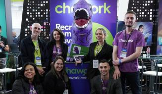 Grupo de sete pessoas da equipe Chawork posa sorridente em um estande de evento. Ao centro, destaca-se um mascote inflável de dinossauro roxo com a boca aberta, posicionado atrás de um balcão com o logotipo da empresa.
