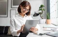 Mulher de óculos e camisa branca sorri ao usar um tablet em um escritório moderno e bem iluminado.