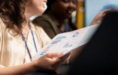 Duas mulheres em ambiente corporativo analisam juntas um relatório com gráficos coloridos.