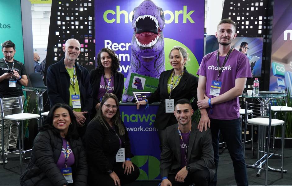 Grupo de sete pessoas da equipe Chawork posa sorridente em um estande de evento. Ao centro, destaca-se um mascote inflável de dinossauro roxo com a boca aberta, posicionado atrás de um balcão com o logotipo da empresa.