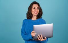 Uma jovem de blusa de tricô azul segura um notebook e olha para o lado com um sorriso alegre, sobre fundo azul.