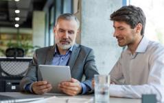 Dois homens de negócios analisam juntos um tablet em uma reunião, sugerindo mentoria ou discussão de resultados.