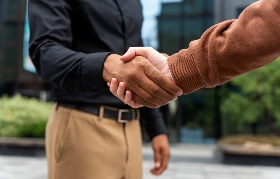 Close-up de um aperto de mão firme entre dois profissionais em um ambiente externo; um veste camisa preta e calça bege, enquanto o outro usa um blusão marrom, simbolizando um acordo ou parceria.