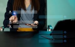 Profissional em escritório usando um tablet com interface de recrutamento/contatos sobreposta, com laptop aberto na mesa ao lado.