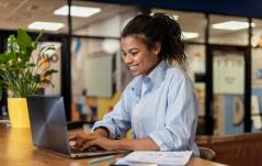Jovem profissional sorridente de camisa azul trabalha em um notebook em um escritório moderno, com uma planta decorativa e documentos à sua frente.