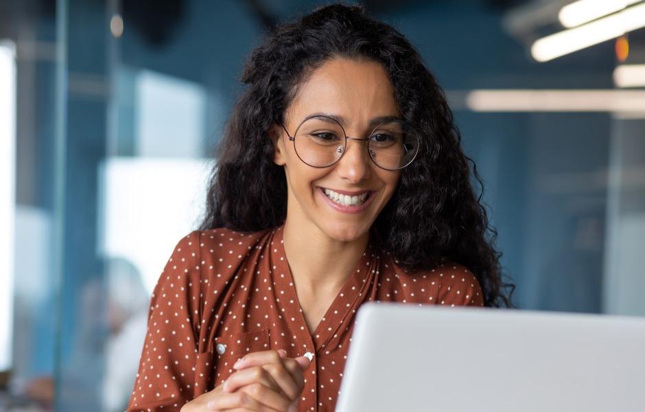 Mulher sorridente usando óculos enquanto olha para o notebook, demonstrando entusiasmo e engajamento no trabalho.