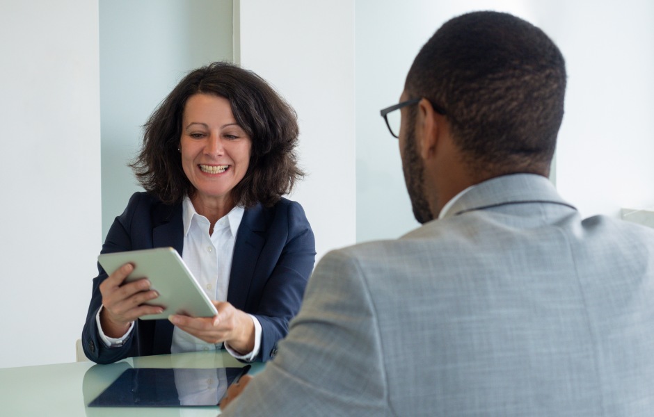 Uma executiva sorridente mostra o conteúdo de um tablet para um colega sentado à sua frente, em um ambiente corporativo iluminado e minimalista.