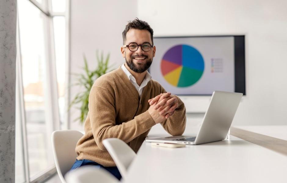 Homem de óculos sorrindo à mesa com notebook, tendo ao fundo um gráfico de pizza colorido projetado na parede.