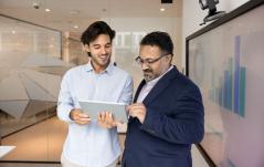 Dois homens de traje social discutem dados exibidos em um tablet em ambiente de escritório moderno, promovendo comunicação eficiente e análise de negócios.