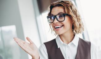 Mulher empresária sorridente usando óculos de grau, vestindo camisa branca e colete, em ambiente de escritório, demonstrando entusiasmo e confiança.