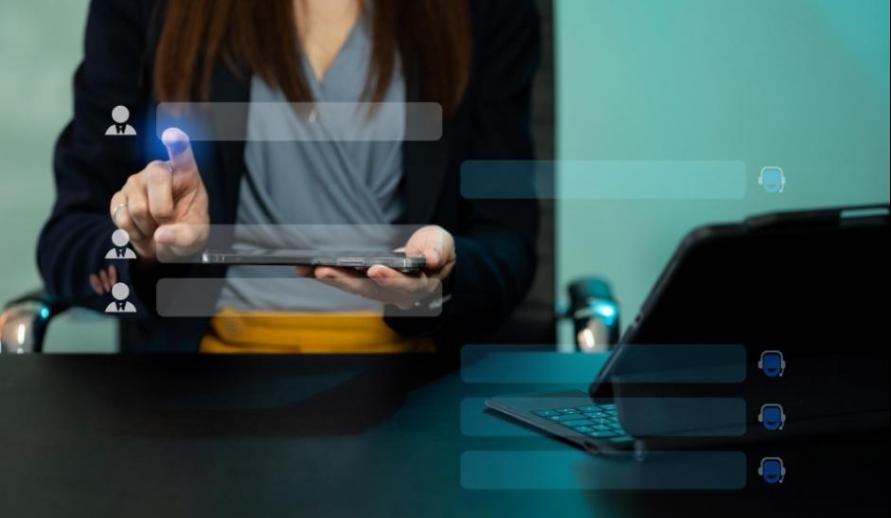 Profissional em escritório usando um tablet com interface de recrutamento/contatos sobreposta, com laptop aberto na mesa ao lado.