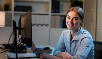 Mulher sorridente usando computador em escritório moderno, trabalhando de casa ou em ambiente profissional, representando produtividade e tecnologia.