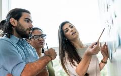 Três profissionais analisam documentos fixados na parede, em uma sessão de brainstorming ou revisão de projeto.
