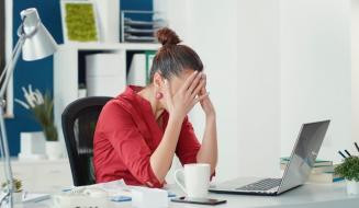Mulher sentada em uma mesa de escritório com as mãos cobrindo o rosto, demonstrando cansaço ou estresse diante de um notebook aberto e uma xícara de café.