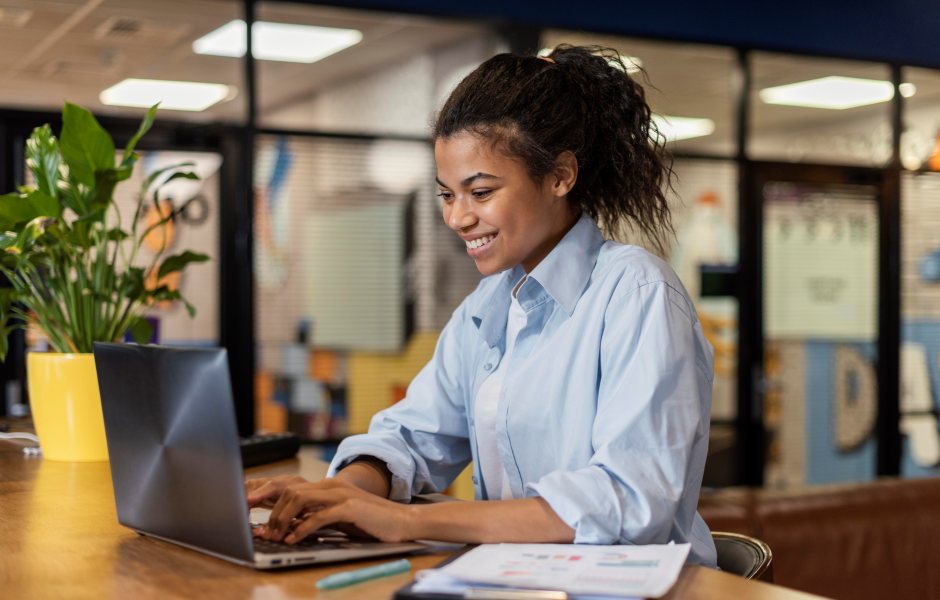 Jovem profissional sorridente de camisa azul trabalha em um notebook em um escritório moderno, com uma planta decorativa e documentos à sua frente.