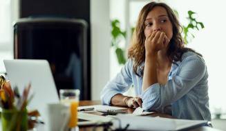 Mulher com expressão preocupada e pensativa diante do notebook, com calculadora e documentos à mesa, sugerindo ansiedade financeira.
