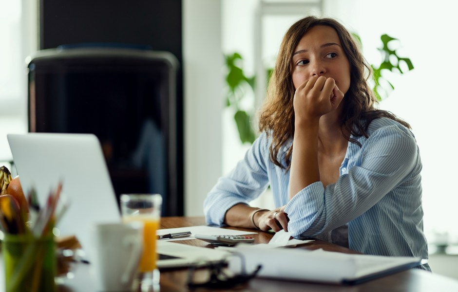 Mulher com expressão preocupada e pensativa diante do notebook, com calculadora e documentos à mesa, sugerindo ansiedade financeira.