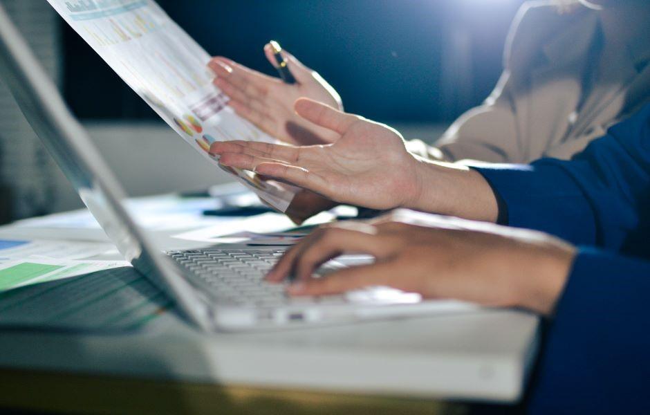 Duas pessoas trabalham juntas, uma digitando no notebook enquanto a outra segura um relatório com gráficos.