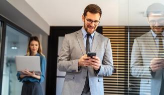 Homem de terno sorrindo ao mexer no celular, com colega ao fundo segurando um notebook.