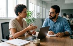 Mulher de cabelo curto e enrolado gesticula com as mãos enquanto conversa com um colega homem de óculos e barba em uma mesa de trabalho. O homem escuta atentamente com uma caneta na mão sobre um caderno, e entre eles estão um notebook aberto, uma xícara de café e relatórios impressos.