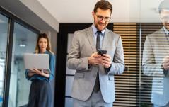 Homem de terno sorrindo ao mexer no celular, com colega ao fundo segurando um notebook.