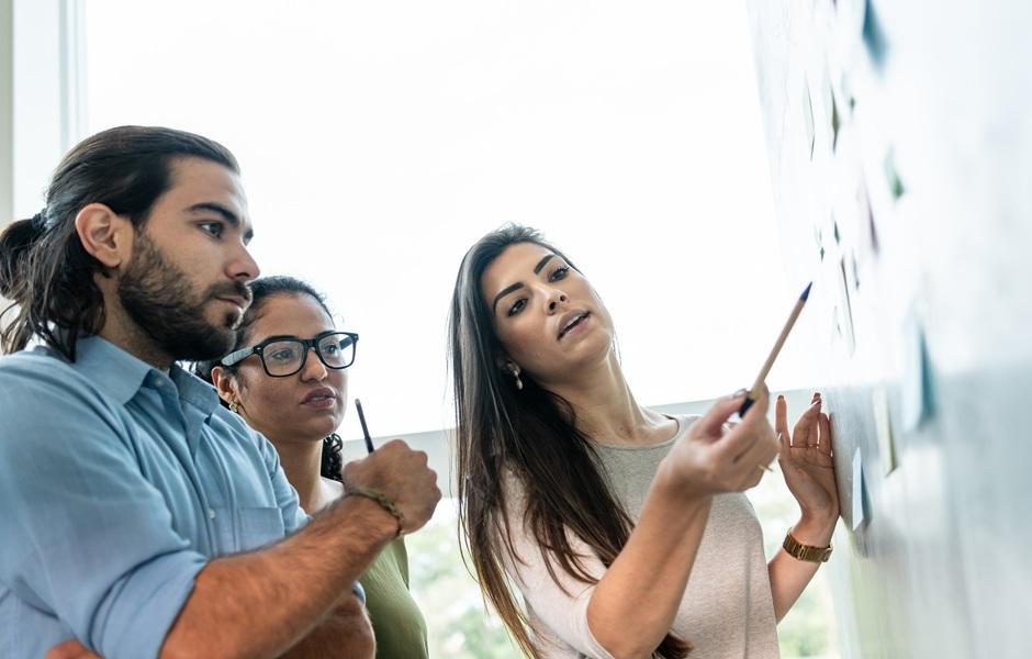 Três profissionais analisam documentos fixados na parede, em uma sessão de brainstorming ou revisão de projeto.