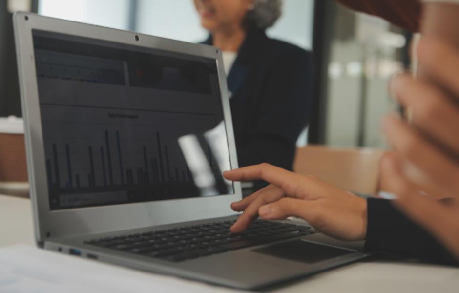 Mãos digitando em notebook com gráfico de barras na tela, em ambiente de reunião corporativa.