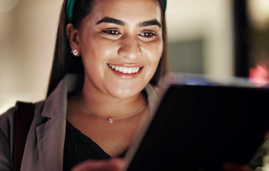 Jovem mulher sorrindo enquanto usa um tablet, com a luz da tela iluminando seu rosto.
