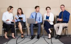 Cinco profissionais sentados em cadeiras de uma sala de espera, com laptops e pastas, demonstrando diferentes expressões de ansiedade ou conversa.